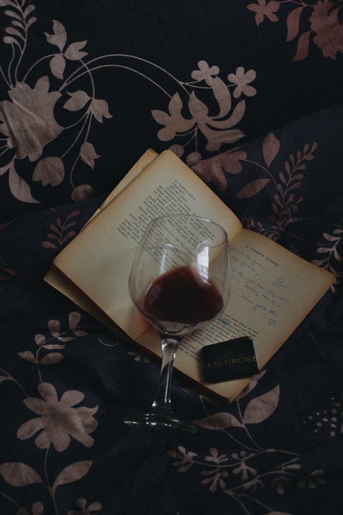 gallery-4 Open vintage book with a glass of red wine resting on floral fabric, evoking elegance and nostalgia.