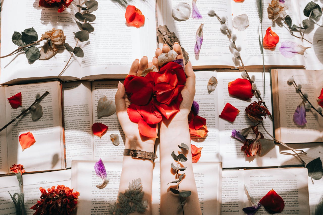 gallery-3 Hands with red petals over open books, surrounded by scattered flowers and petals for artistic composition.